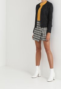 Giacca nera testurizzata, maglione senape, gonna a quadri e stivaletti bianchi con tacco. L'outfit mette in mostra stili a strati e colori a contrasto.