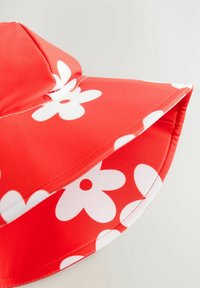 Cappello a secchiello rosso realizzato in tessuto liscio, caratterizzato da grandi motivi floreali bianchi. La visiera è ampia e arrotondata con cuciture a contrasto.