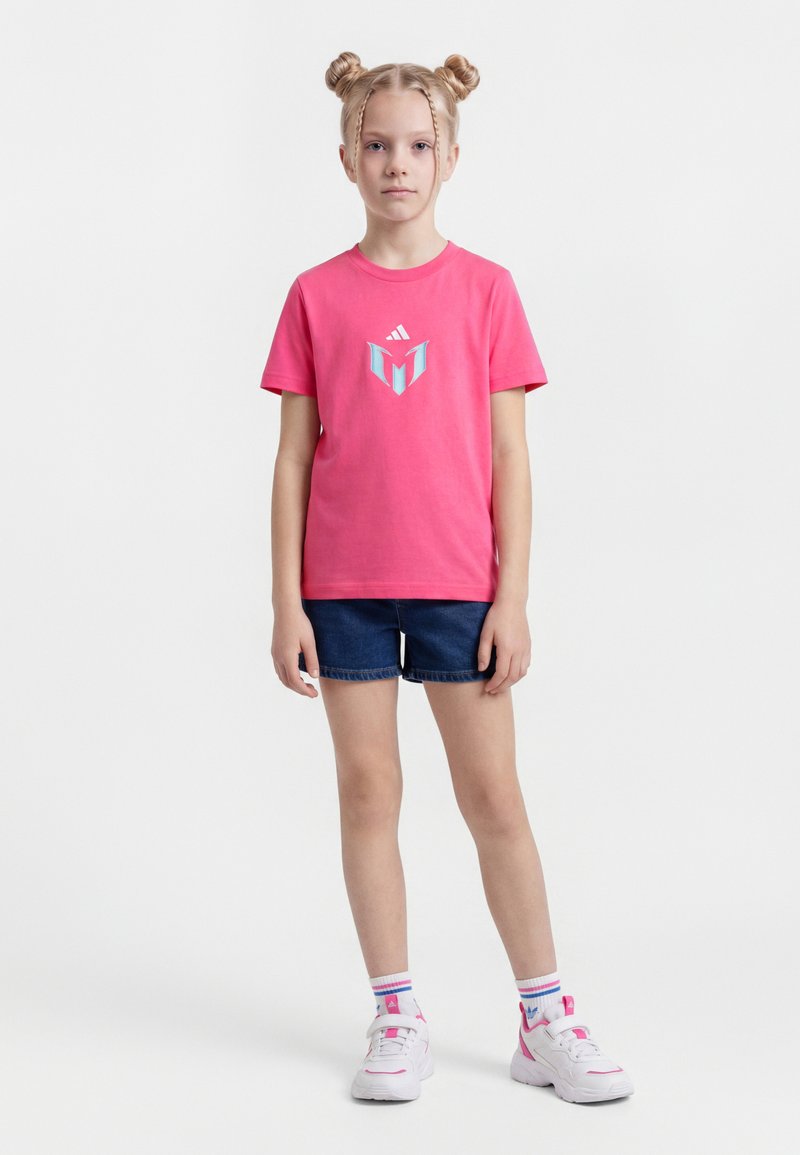 Νεαρή κοπέλα με δύο κότσους, που φοράει ροζ μπλούζα Adidas, σκούρα μπλε σορτς, λευκές κάλτσες Adidas και λευκά αθλητικά παπούτσια, στέκεται μπροστά σε λευκό φόντο.