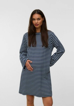 Femme aux longs cheveux foncés portant une robe à manches longues rayée bleu marine et blanc, debout avec une main sur la taille et l'autre sur le ventre.