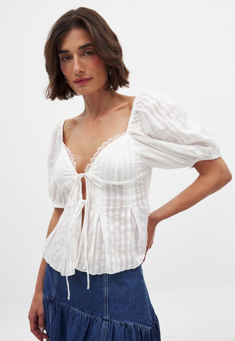 Weißes Bluse mit Puffärmeln, Spitzendetails und Knüpfverschluss, mit vertikaler gestreifter Textur und seitlichen Schlitzen. Kombiniert mit einem Jeansrock.