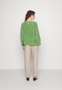 Blusa verde, texturizada, com mangas longas e um pequeno detalhe em forma de buraco na parte de trás, combinada com calças bege de pernas retas e sandálias pretas de salto bloco.