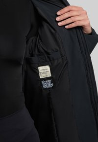 Sort isoleret jakke med en glat foring, der har indvendige lommer og mærker. Designet inkluderer en lynlås og syninger.