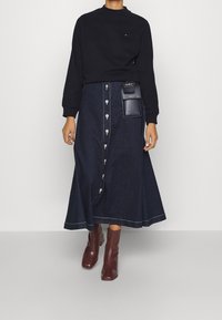 Czarny sweatshirt z okrągłym dekoltem noszony na długiej dżinsowej spódnicy z przednimi guzikami, w ciemnoniebieskim odcieniu, z boczną kieszenią z metalowymi akcentami.