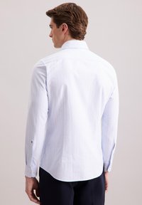Uomo che indossa una camicia a righe sottili blu chiaro con maniche lunghe, in piedi con la schiena verso la macchina fotografica su uno sfondo chiaro e semplice.