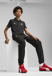 Camiseta polo negra con el logo de Ferrari, pantalones deportivos negros con el logo y zapatillas rojas de Puma. Diseño sencillo, tejido ligero y corte atlético.