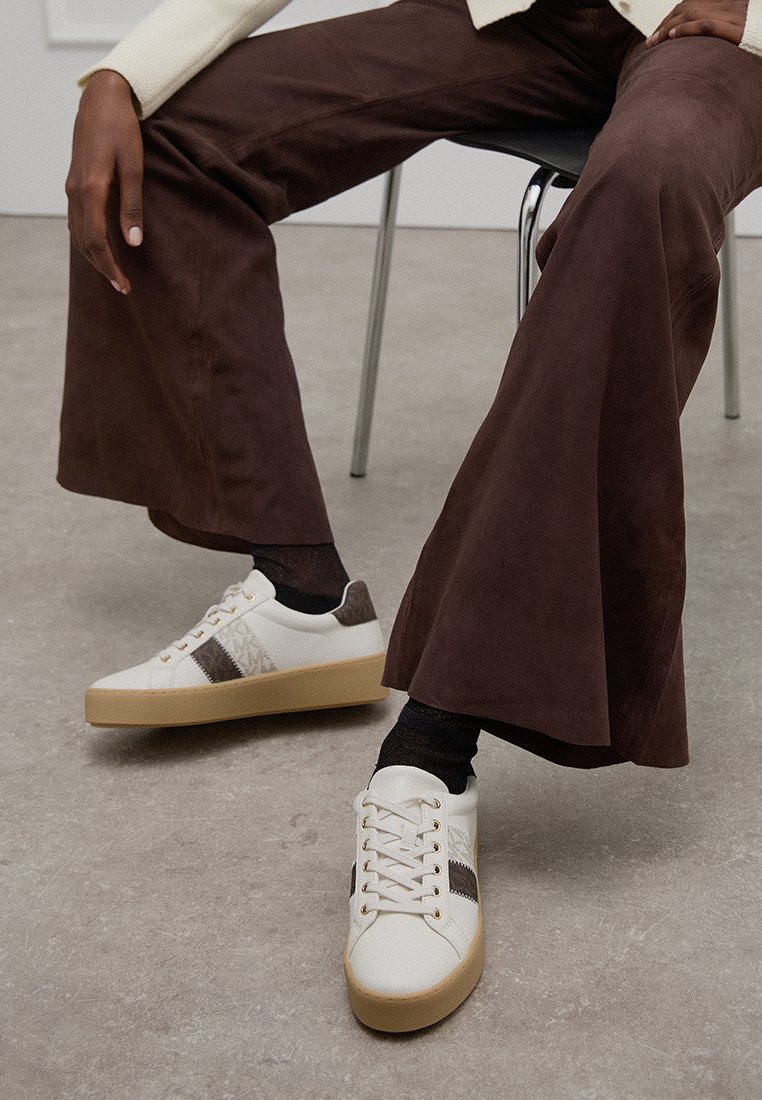 Zapatillas blancas con acentos marrones y un panel texturizado, combinadas con pantalones de pierna ancha marrones oscuros, sobre un fondo neutral.