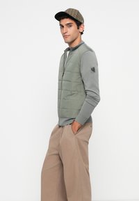 Young man wearing a green quilted zip-up jacket, gray sleeves, beige wide-leg pants, and a checkered flat cap, standing with hands in pockets.