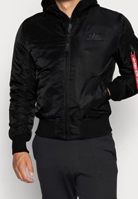 Giubbotto bomber nero con zip frontale, cappuccio, polsini e orlo a costine, tasca laterale e una fascia rossa a contrasto. Materiale liscio e lucido.