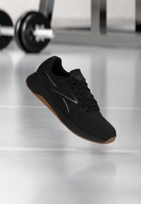 Μαύρο αθλητικό παπούτσι Reebok με σόλα από καουτσούκ, αιωρούμενο στον αέρα πάνω στο πάτωμα του γυμναστηρίου, με θαμπά όργανα άρσης βαρών στο φόντο.