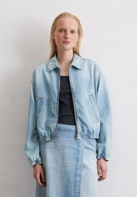 Eine Person mit langen blonden Haaren posiert vor einer schlichten weißen Wand, trägt eine helle Jeansjacke und ein dunkles Oberteil und zeigt ein lässiges Outfit.