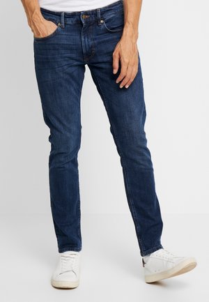 Uomo che indossa jeans slim fit blu scuro e scarpe da ginnastica bianche con una mano in tasca su sfondo uniforme.