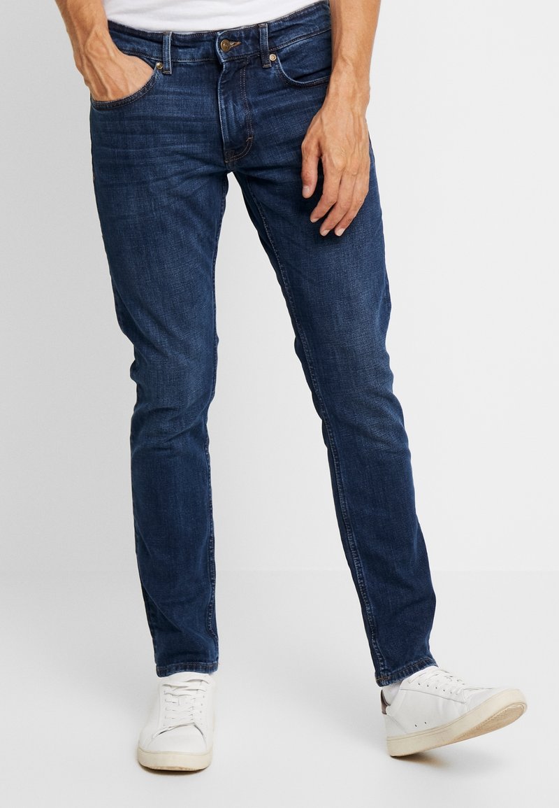 Homme portant un jean slim bleu foncé et des baskets blanches, une main dans la poche, sur fond uni.