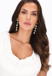 Bijuterii care includ cercei lungi, cu flori, accentuați cu detalii clare și aurii. Model purtând un top alb, texturat, cu un decolteu volan.