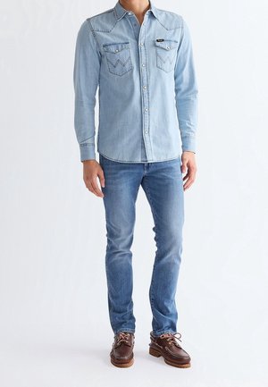 Mann trägt hellblaues Jeanshemd mit Knöpfen, blaue Jeans und braune Lederschnürschuhe, steht vor weißem Hintergrund.