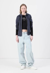 Marineglidet jakke over en svart cropped topp, lyseblå vide jeans og hvite sneakers. Outfiten har subtile logodetaljer.