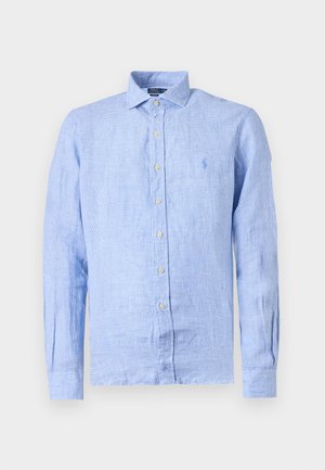 Νέος άντρας στέκεται φορώντας ένα ανοιχτό μπλε πουκάμισο με μακριά μανίκια και κουμπιά, μαύρο παντελόνι, μπεζ μοκασίνια, με το ένα χέρι στην τσέπη, μπροστά σε απλό φόντο.