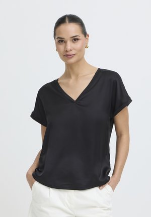 Femme aux cheveux attachés portant un haut noir à manches courtes avec un col en V et un pantalon blanc, mains dans les poches, sur un fond uni.