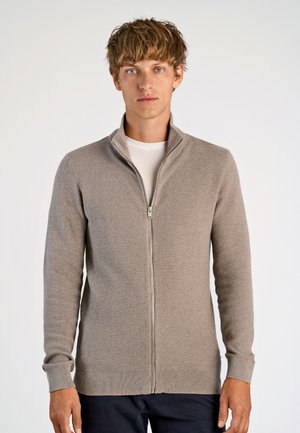 Beige zip-up tröja med texturerad stickning, hög krage och ribbade ärmslut. Bärs över en vit t-shirt.