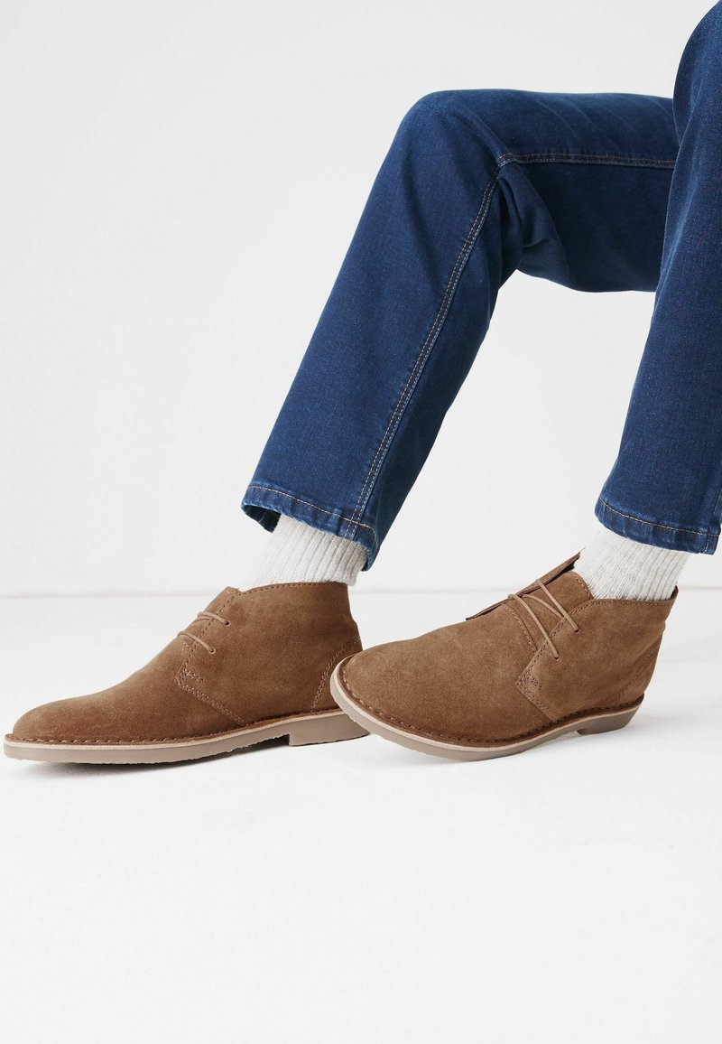 Botas de tobillo de ante marrón con dos agujeros para cordones, suela clara en contraste, usadas con calcetines blancos y pantalones de mezclilla azul.