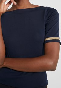 Tmavě modrý top s krátkými rukávy z měkké látky, s širokým výstřihem a zlatými proužky na rukávech.