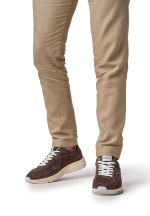 Zapatillas de ante marrón con cordones blancos, con un diseño texturizado y suela de color claro, combinadas con pantalones beige ajustados.