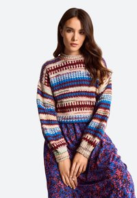 Moteris su ilgais rudais plaukais, vilkinti spalvingą dryžuotą megztinį ir mėlyną bei rožinę raštuotą sijoną, stovi su rankomis sugniaužtomis.