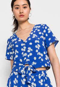 Top cropped azul com mangas curtas, apresentando um padrão de margaridas brancas, decote em V, botões na frente e um detalhe amarrado na cintura.