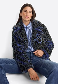Manteau en fausse fourrure imprimé léopard dans des tons sombres avec des accents bleus. Dispose d'un large col et d'un design surdimensionné. Porté sur une chemise rayée et un jean.