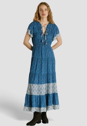 Junge Frau in einem blauen Maxikleid mit Muster, Quastenbändern und weißem gemustertem Saum, kombiniert mit schwarzen Stiefeletten, steht mit den Händen hinter dem Rücken.