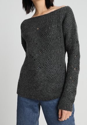 Femme portant un pull en tricot gris foncé avec un col bateau et de légers trous usés, associé à un jean bleu, fond neutre.