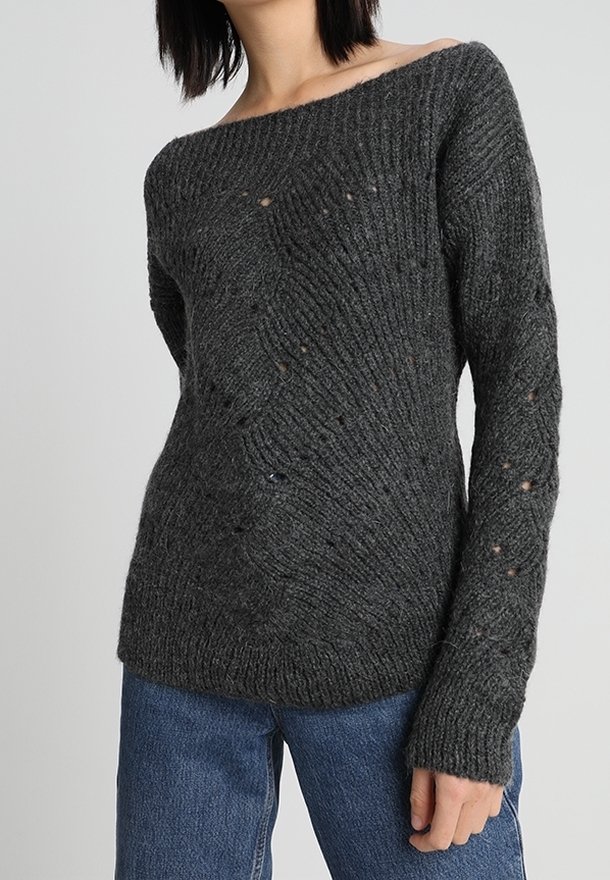 Femme portant un pull en tricot gris foncé avec un col bateau et de légers trous usés, associé à un jean bleu, fond neutre.