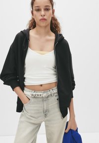 Zwarte ritshoodie over een witte tanktop, gecombineerd met lichte spijkerbroeken en een zilverkleurige gesp-band. Blauwe stof over een arm gedrapeerd.