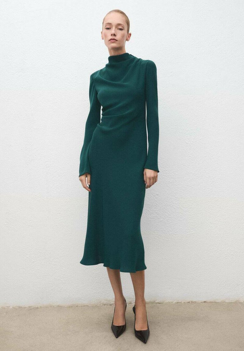 Vestido de manga larga y cuello alto en verde oscuro, hecho de una tela suave, con una silueta ajustada y un dobladillo acampanado, combinado con tacones negros de punta.