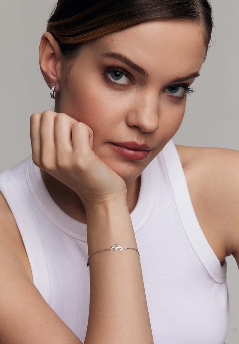Srebrna bransoletka z projektem liścia na delikatnym łańcuszku, z małym akcentem w kolorze turkusowym, noszona na nadgarstku na jasnoszarym tle.