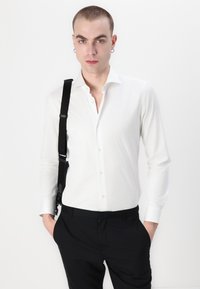 Weißes Langarmhemd mit slim Fit, Button-Down-Kragen und glatter Textur, kombiniert mit schwarzen Hosen und ausgestattet mit einem schwarzen Schultertragegurt.