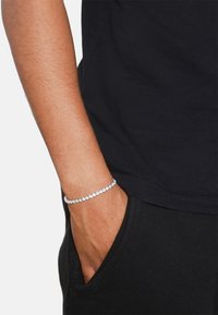 Bracciale d'argento con pietre trasparenti, indossato su un polso. La mano è infilata in una tasca, con una camicia nera visibile. Texture liscia e forma rotonda.