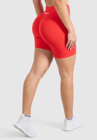 Rode high-waisted shorts gemaakt van rekbaar materiaal, met een gladde textuur en een logo op de tailleband. Gedragen met witte sportieve schoenen.