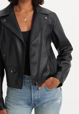 Femme portant une veste en cuir noire de motard par-dessus un haut noir avec un jean bleu clair, collier en or et bracelets, main dans la poche.