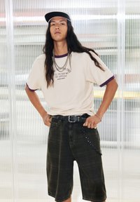 Beige t-shirt met paarse accenten en tekst, gecombineerd met zwarte denim shorts met een kettingdetail. Model heeft lang haar en draagt een pet.