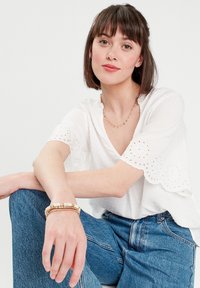 White blouse with eyelet detailing at sleeves, paired with blue high-waisted jeans. Accessories include a layered gold necklace and a beaded bracelet.