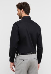 Camicia nera a maniche lunghe con colletto standard, con polsini abbottonati. Pantaloni grigi con tasche posteriori e bottoni visibili, cintura inclusa.