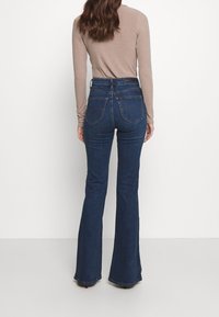 Džínsy s vysokým pasem a rozšířenými nohavicemi v tmavě modré denimové barvě, s kapsami a jemnou texturou. Spárováno s pleteným topem s dlouhým rukávem v taupe barvě.