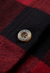 Tessuto a quadri rosso e nero con un grande bottone beige con la scritta "SUPERDRY" incisa. Texture morbida con dettagli di cucitura visibili.
