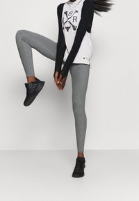Šedé legíny v kombinaci s bílým a černým dlouhým rukávem s grafickým potiskem. Oblečení doplňují černé sportovní boty.