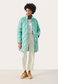 Jachetă matlasată de culoare teal cu guler înalt, fermoar auriu și buzunare laterale, asortată cu pantaloni bej deschis și mocasini maronii.