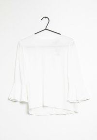 Vit blus med utsvängda ärmar, lättviktsmaterial, rundad halsringning och asymmetrisk fåll. Hänger på en svart krok mot en enfärgad bakgrund.