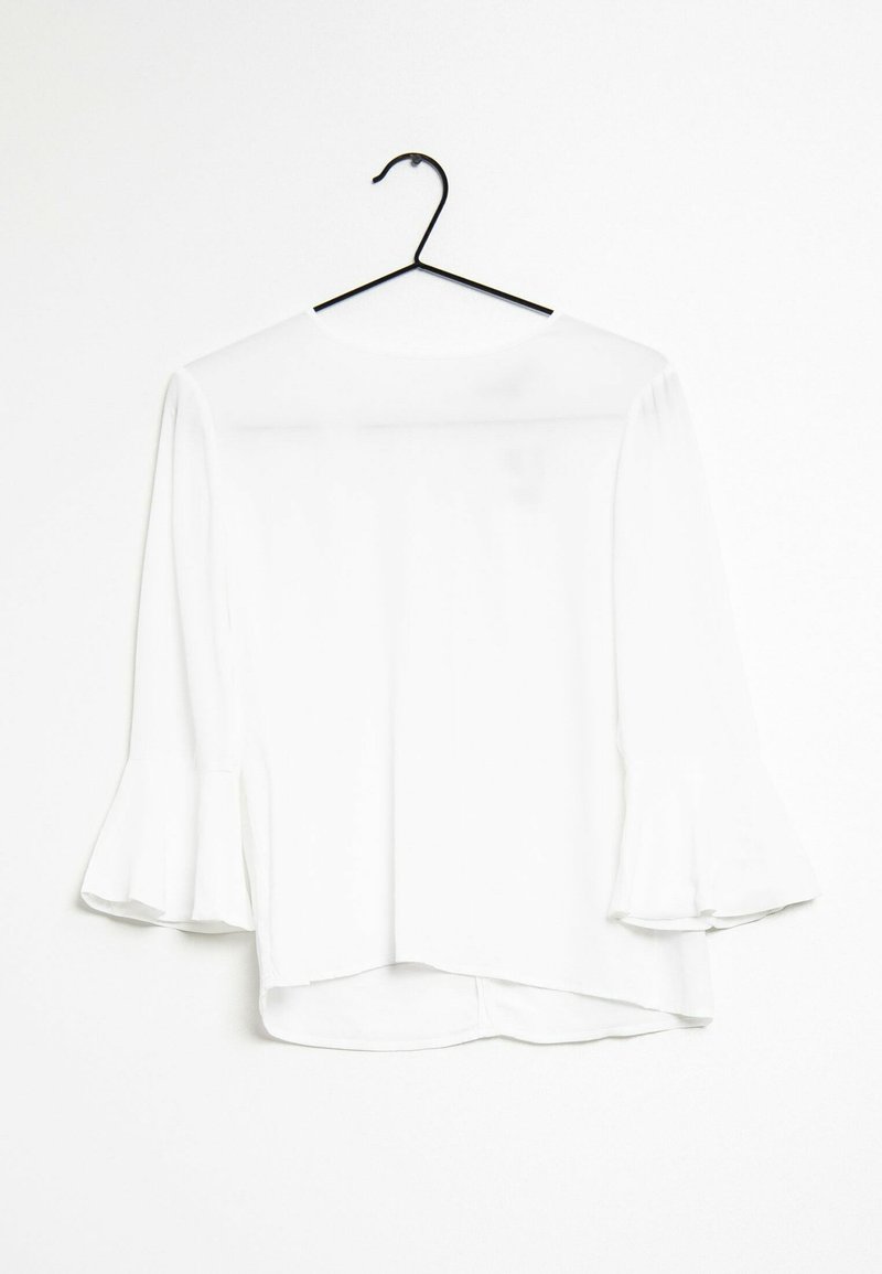 Vit blus med utsvängda ärmar, lättviktsmaterial, rundad halsringning och asymmetrisk fåll. Hänger på en svart krok mot en enfärgad bakgrund.