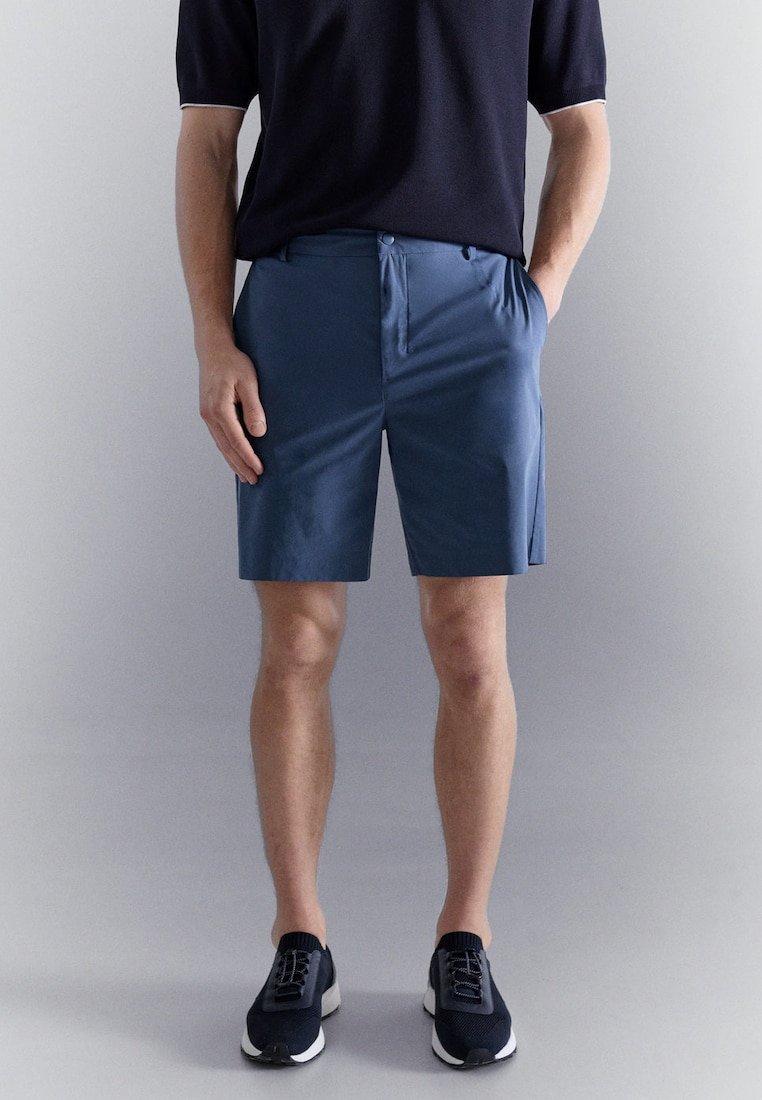 Homme portant un short bleu marine décontracté, une chemise noire à manches courtes et des baskets noires, debout devant un fond gris uni.
