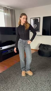 Modello che indossa una camicia nera con bottoni e jeans grigio chiaro, abbinati a calzature color cuoio, su un tappeto grigio testurizzato in una stanza minimalista.